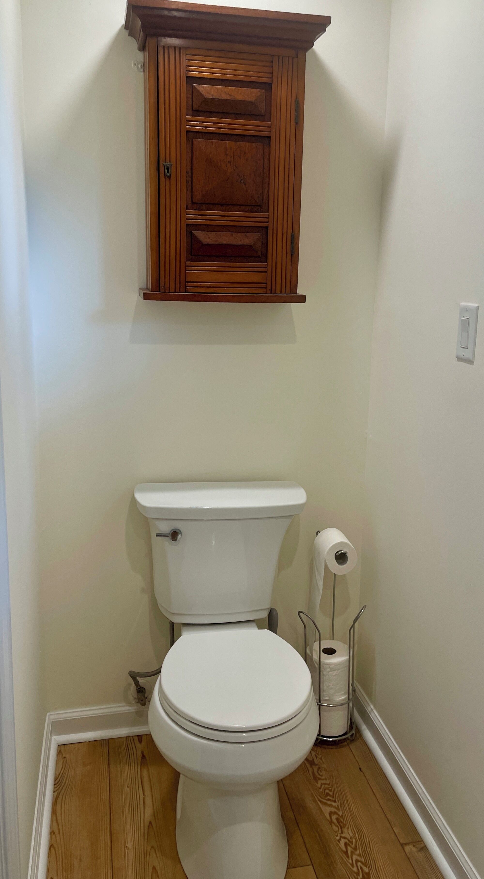 A bathroom with a white toilet, wooden cabinet above, and a toilet paper holder.
