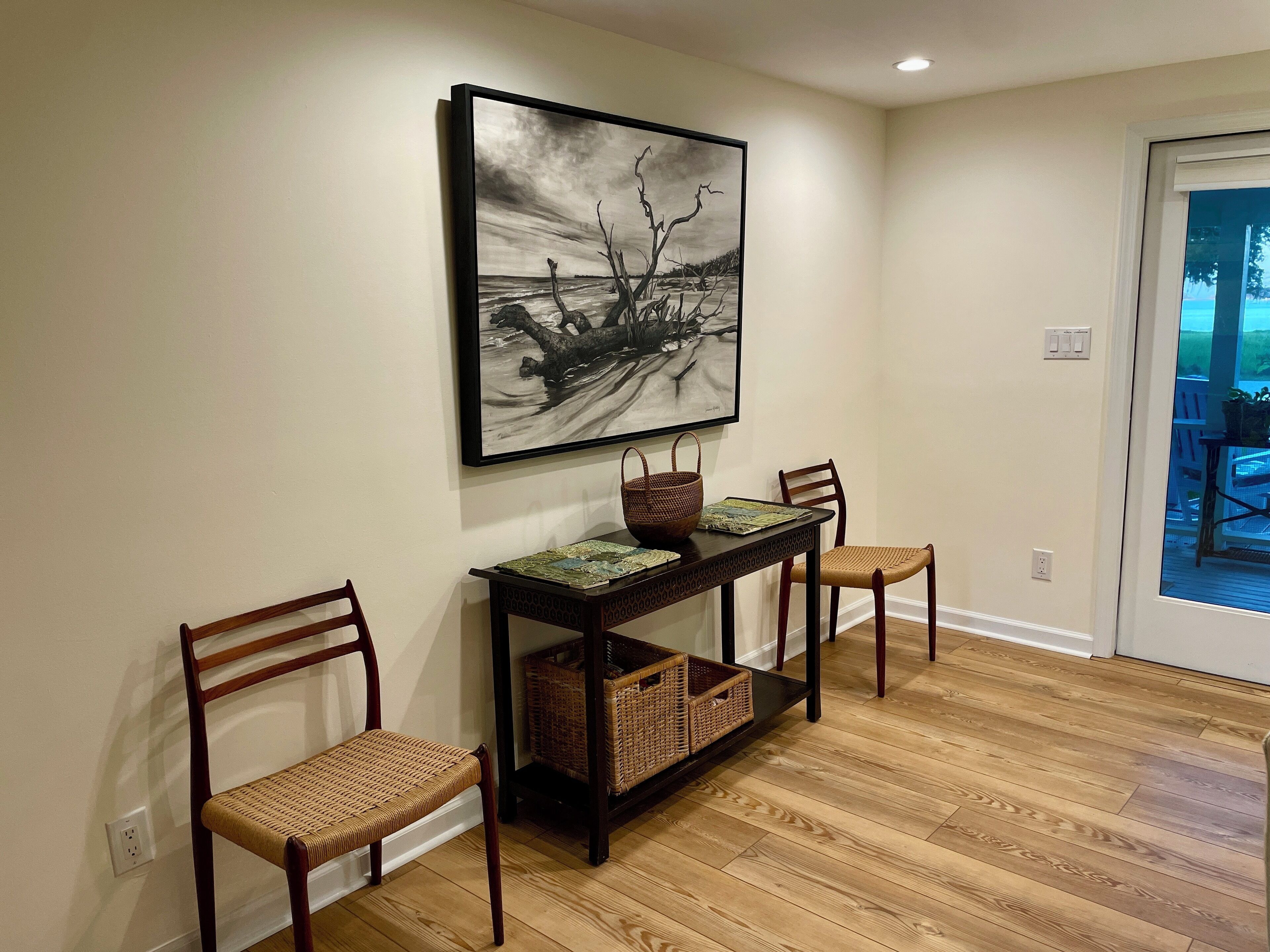 A dining area with a dark wooden table, two wicker chairs, a woven basket, and a framed black and white painting on the wall.