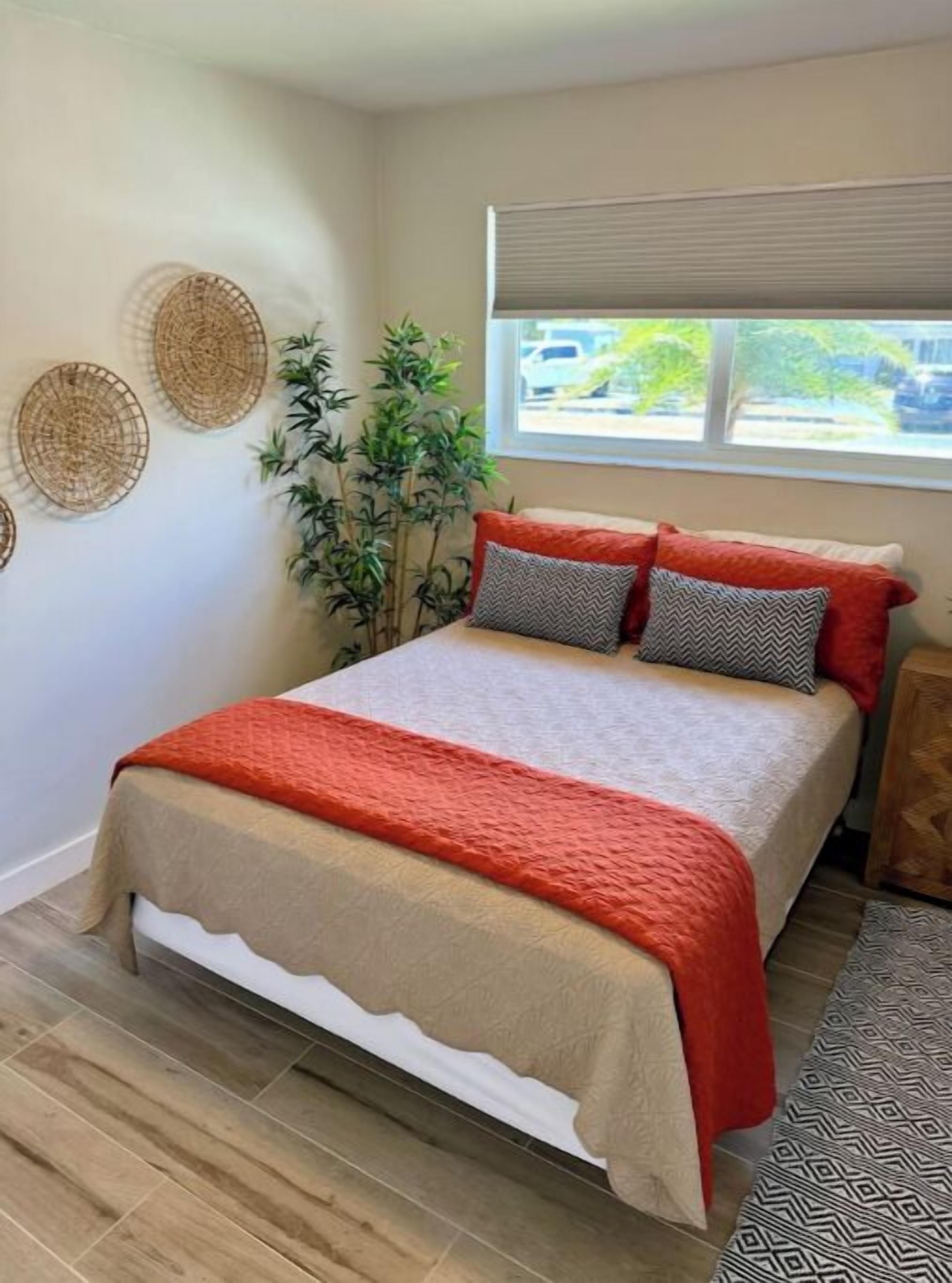 A neatly made bed with a red and beige bedspread, two pillows, and a small plant beside the window.