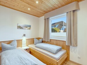 A bedroom with two beds, wooden ceiling, a window with curtains, a wall-mounted light, and a beach-themed picture. - Vacation home Heide by Interhome (Seebad Altefähr)