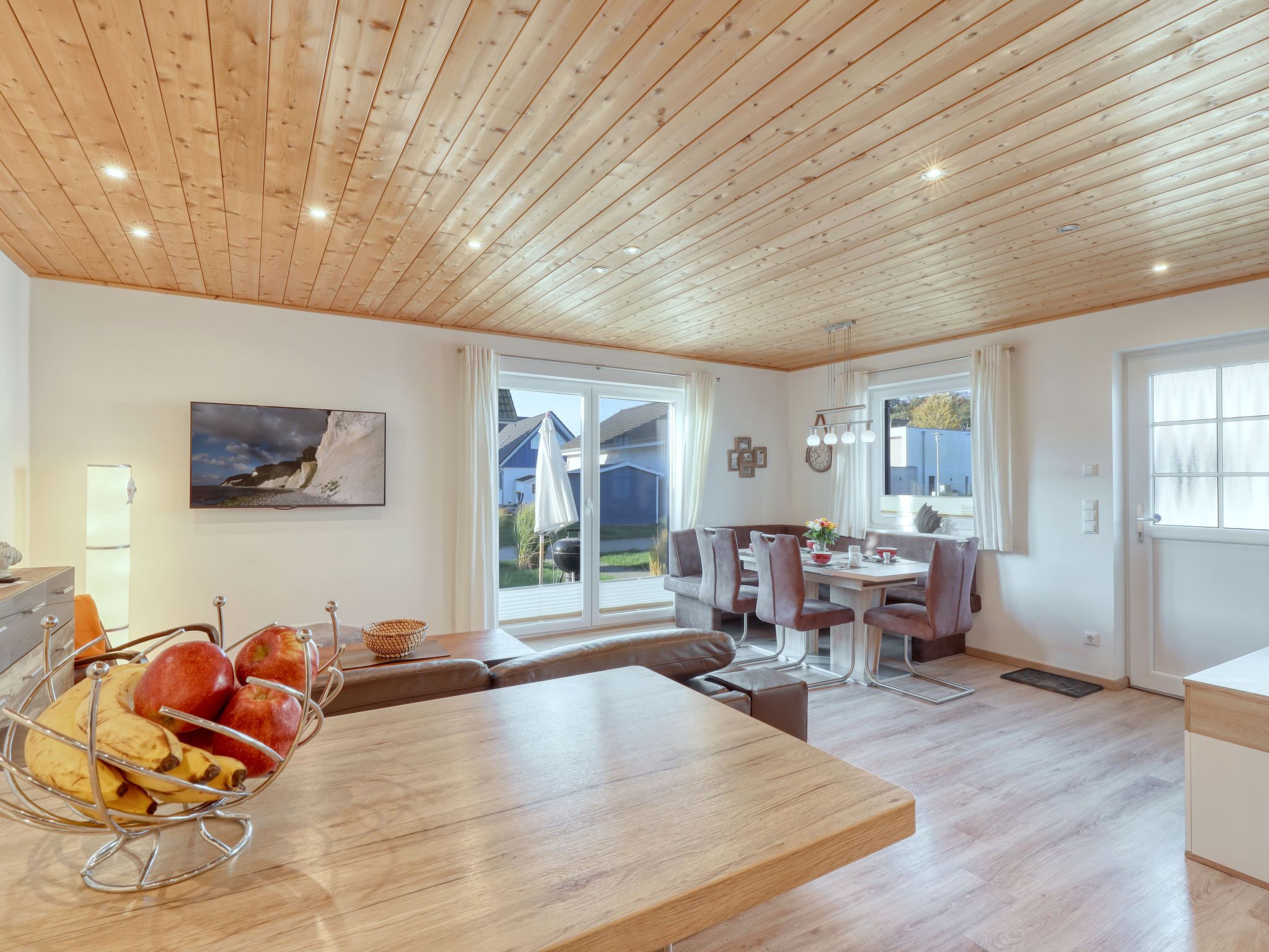 Eine moderne Küche mit Holzboden, ein Essbereich mit Tisch und Stühlen sowie ein Blick ins Freie durch große Fenster.