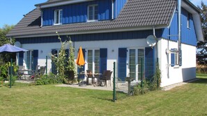 Una casa de dos pisos con techo inclinado, ventanas grandes con contraventanas y un patio vallado con mobiliario exterior.