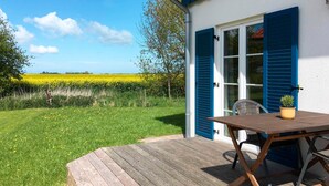 Una terraza de madera con una mesa y sillas, una puerta azul y vistas a un campo amarillo.