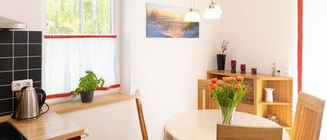 Une cuisine moderne équipée d’une cuisinière, d’un évier et de meubles de rangement rouges. On y trouve également un coin repas avec une table, des chaises et un vase de fleurs. Une fenêtre avec des rideaux et un tableau accroché au mur sont également visibles.