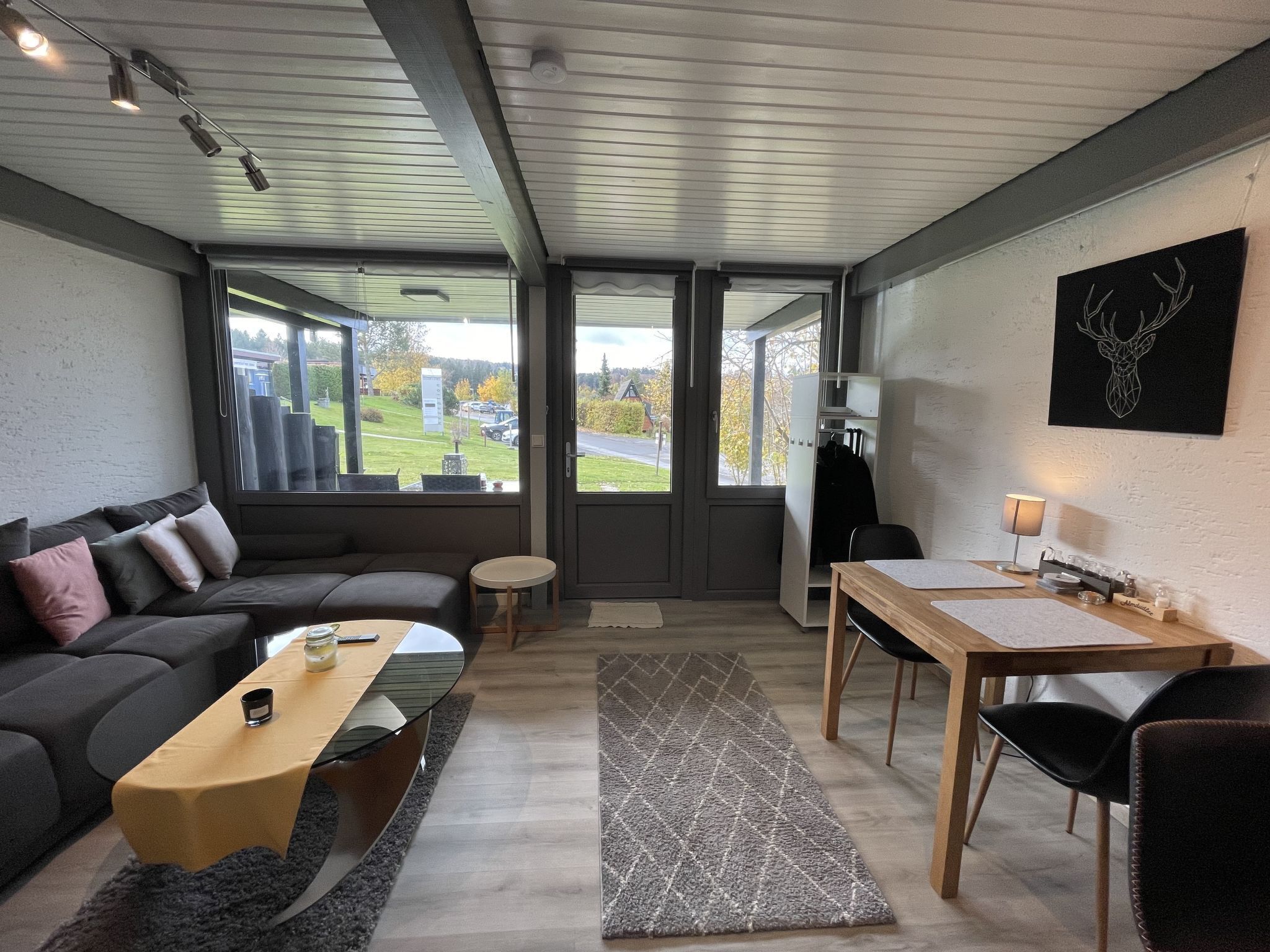 A modern living room with a sofa, coffee table, and a dining area with a table and chairs. There is a large window providing a view of the outdoors.
