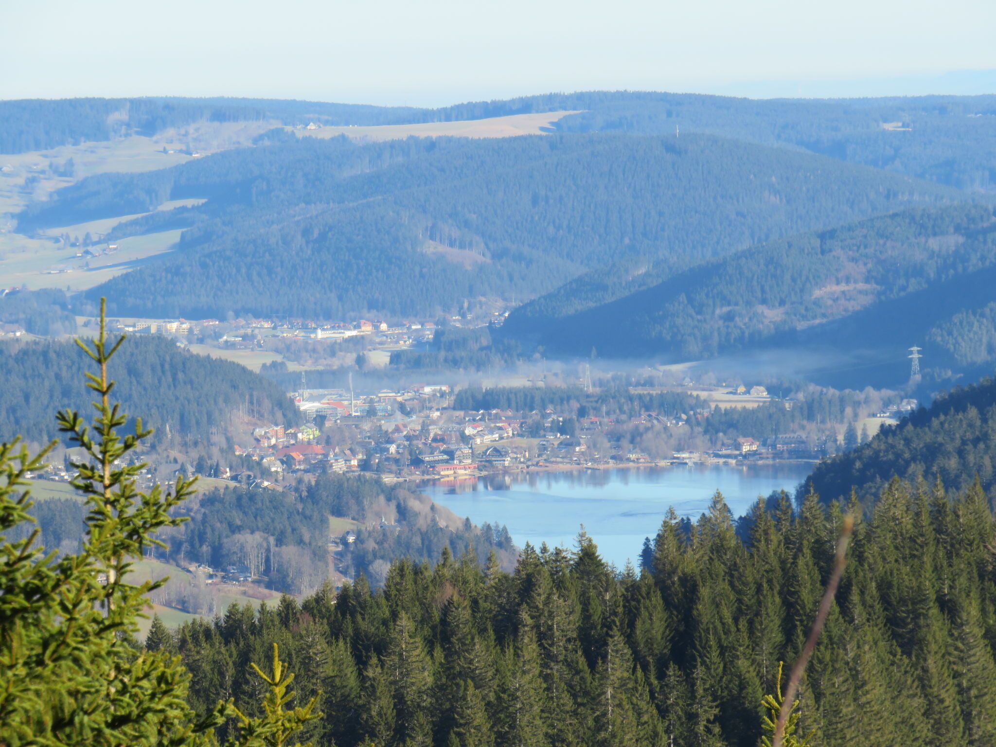 Vista panorámica de un pueblo ubicado en un valle, con un lago en primer plano, rodeado de densos bosques y colinas.
