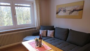 Ein gemütliches Wohnzimmer mit einem dunkelgrauen Sofa, einem Couchtisch mit Flasche und Gläsern und einem Fenster mit Vorhängen.