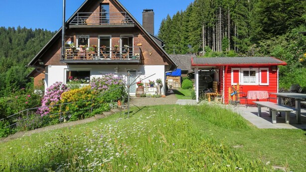 Eine zweistöckige Chalet mit Balkon, umgeben von üppiger Vegetation und einem kleinen roten Gebäude.