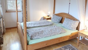 Een houten hemelbed met een groene matras en bedekte kussens, een nachtkastje met een lamp en een raam met gordijnen.