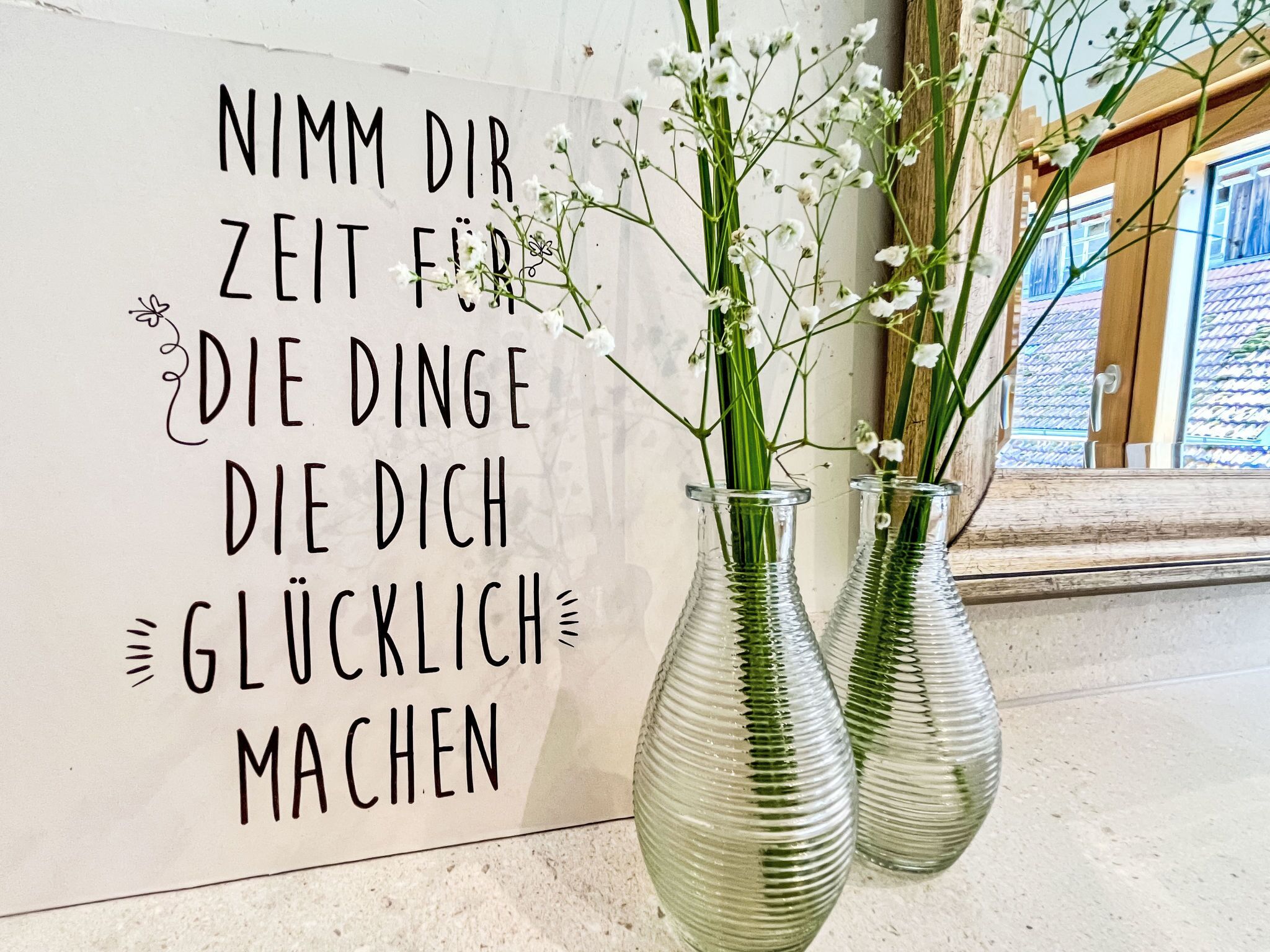 Two glass vases with white flowers on a wall with a German text.