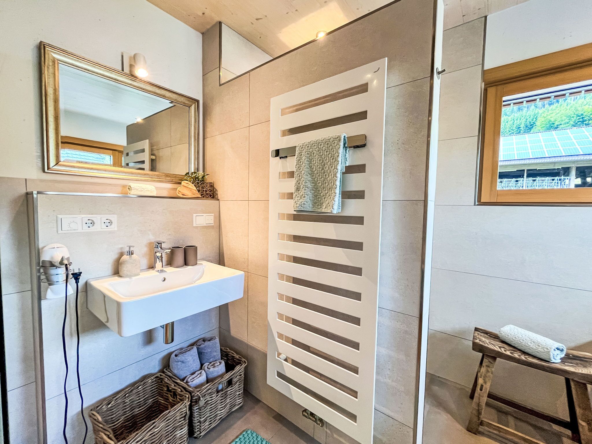 A modern bathroom with a white sink, mirror, and towel rack. There is a window with a view of greenery outside.