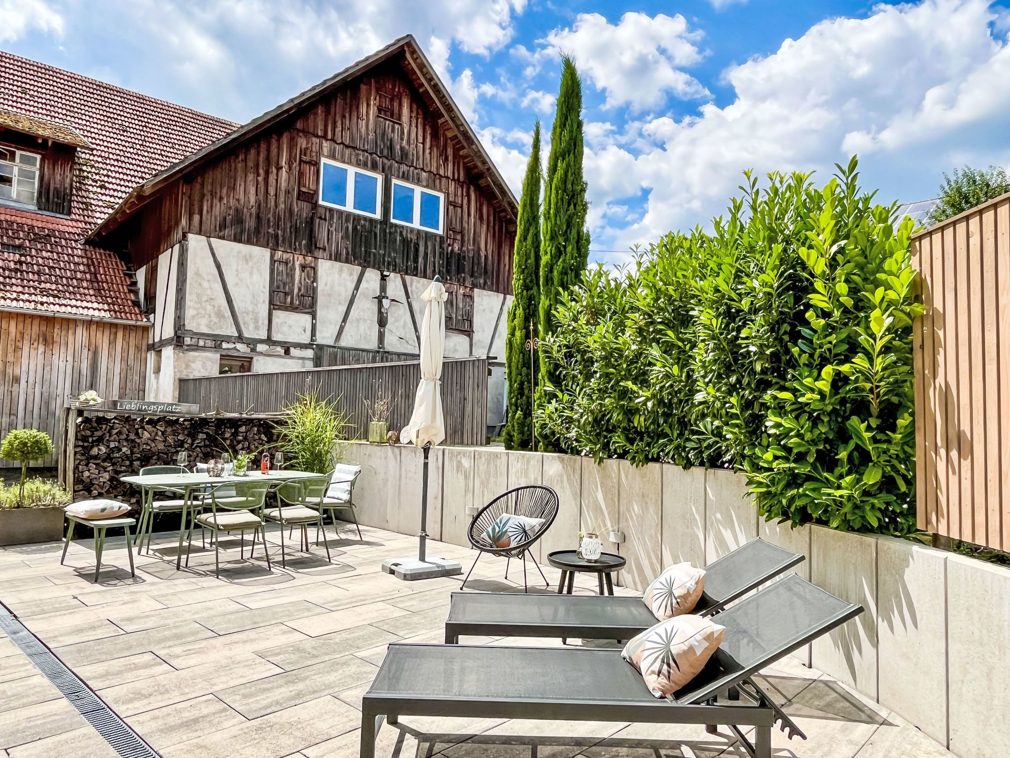 A wooden house with a tiled roof, a patio with outdoor furniture, and a well-maintained hedge.