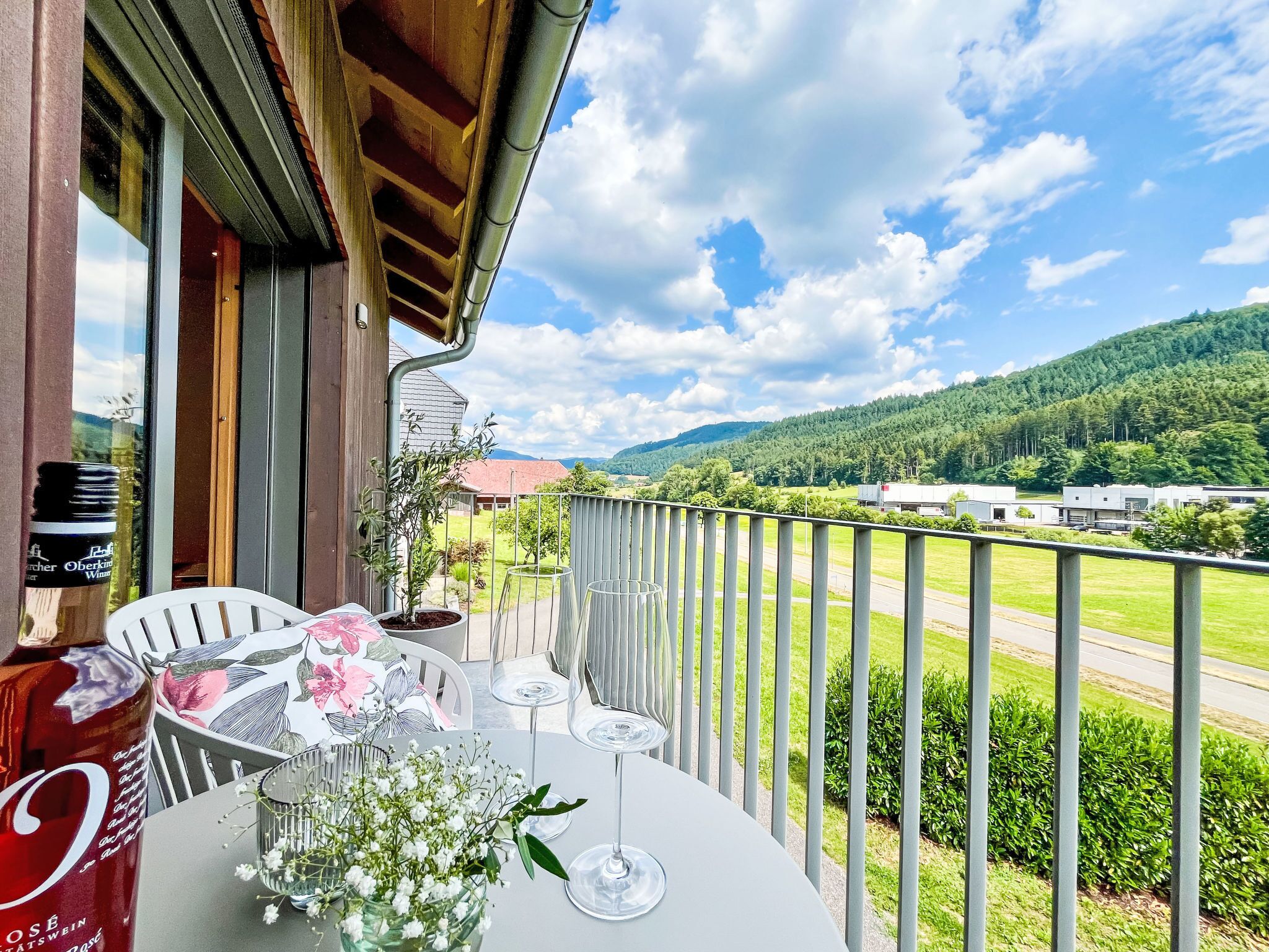A balcony with a table set for two, a bottle of wine, and a potted plant.