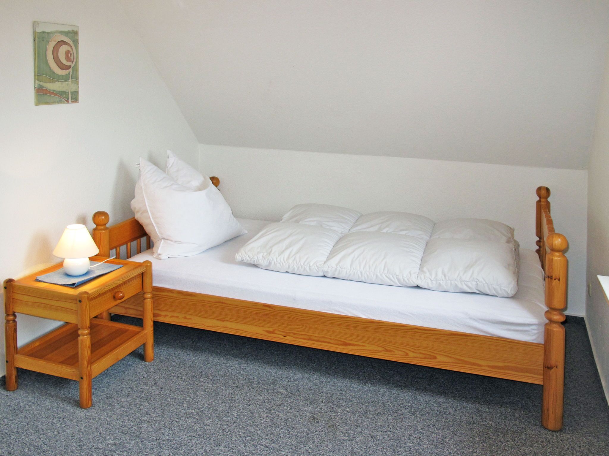 A single bed with a wooden frame and white bedding, a bedside table with a lamp, and a wall-mounted artwork.