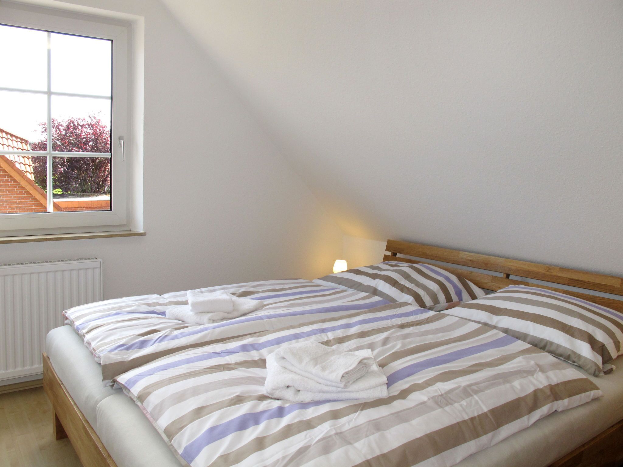 A double bed with striped bedding and two towels, a window with a view of a brick building and trees, and a bedside lamp.