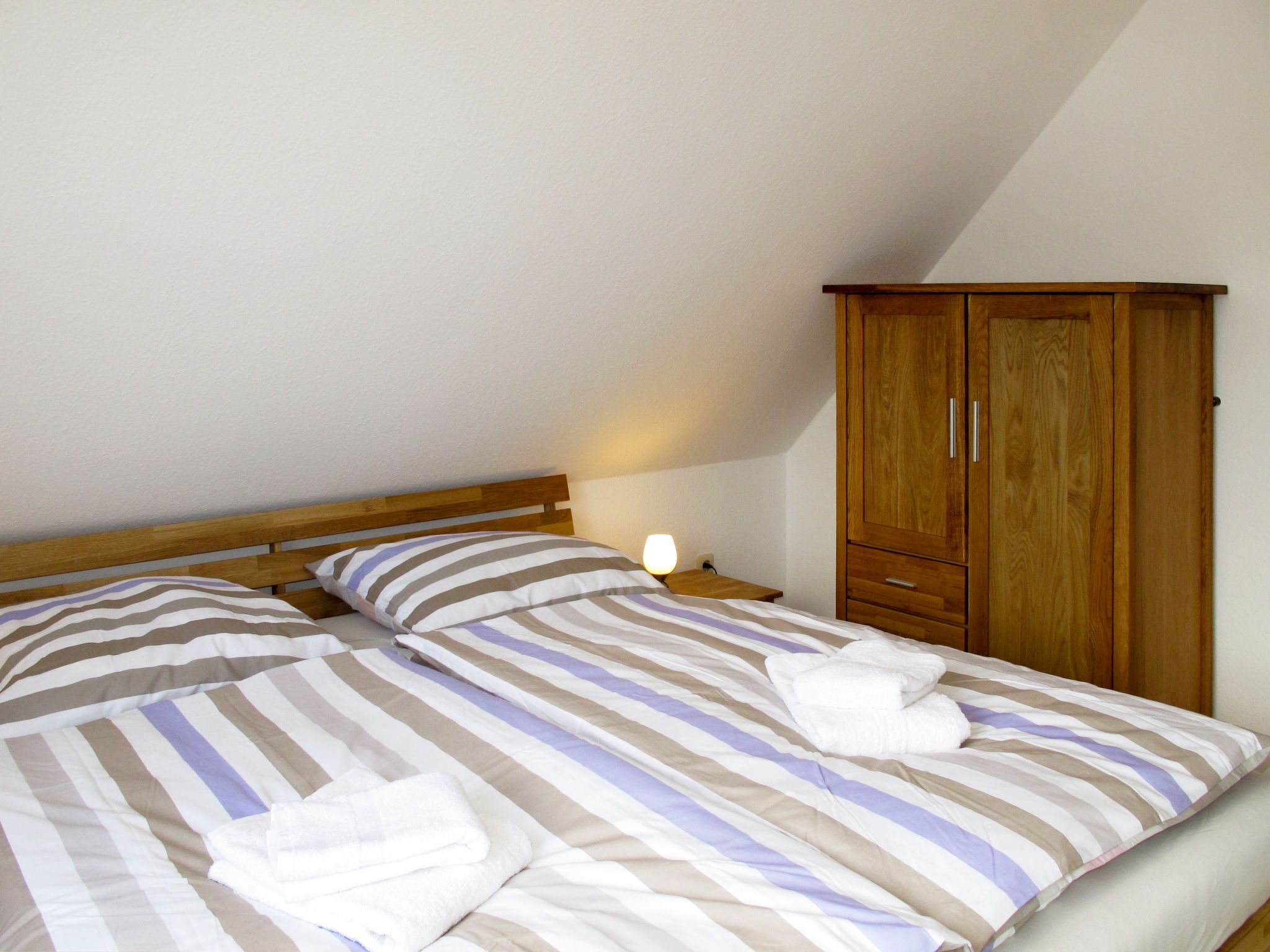 A bedroom with two beds, wooden headboards, a nightstand with a lamp, and a wooden wardrobe.