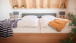 A bedroom with a wooden ceiling, two bedside tables with lamps, a bed with white bedding, and a shelf above the bed with books and plants.