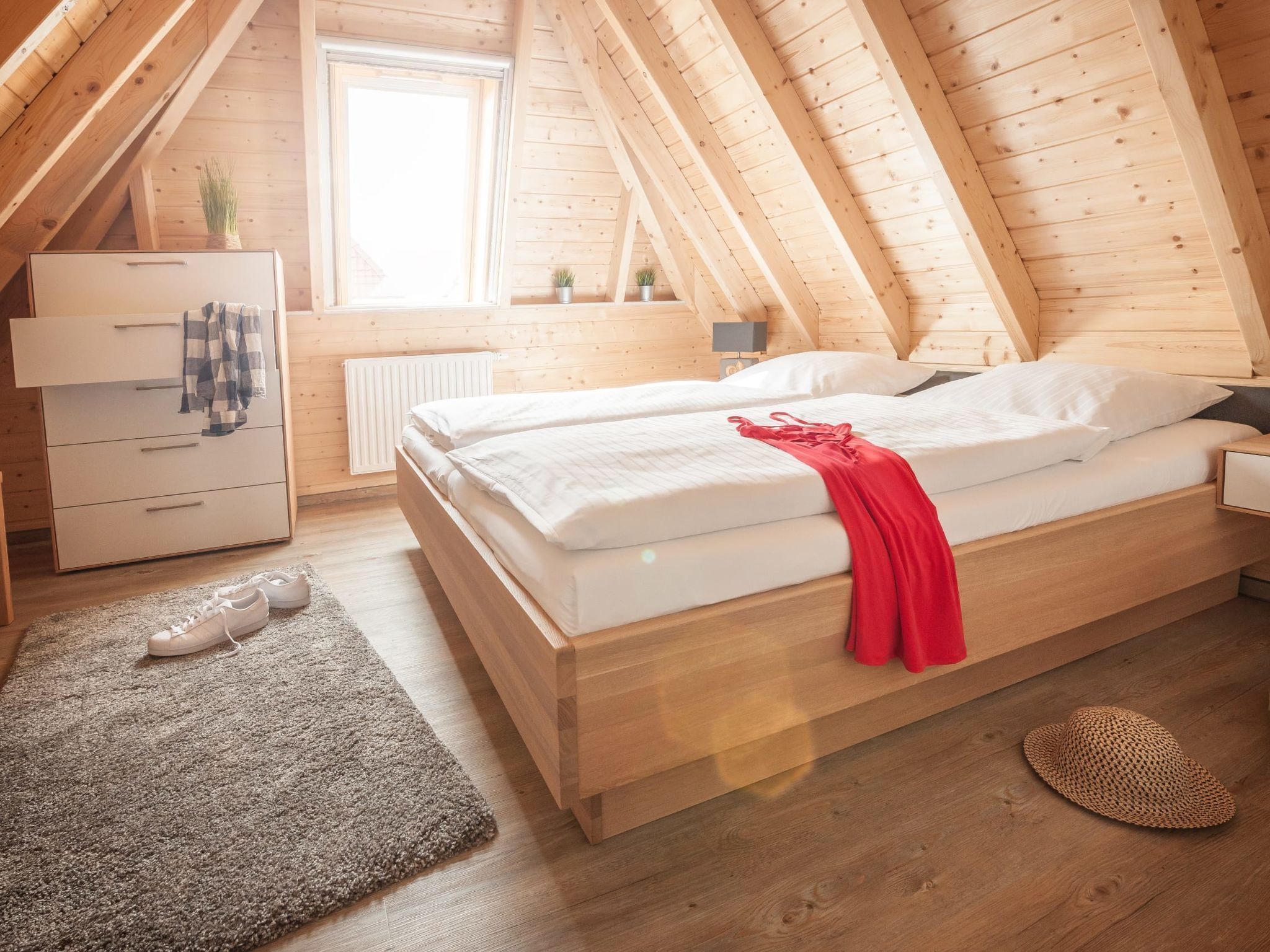 Une chambre avec un plafond en bois, un lit avec une literie blanche, une couverture rouge, une commode, un radiateur et une fenêtre.