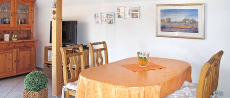 Ein Essbereich mit einem runden Tisch, der mit einer orangefarbenen Tischdecke bedeckt ist, Holzsesseln, einem kleinen Topfpflanzen und einer Uhr an der Wand.