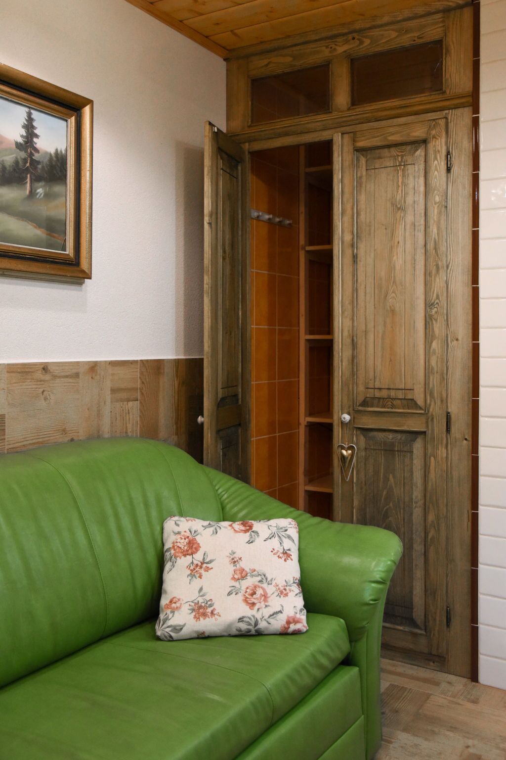 Un canapé en cuir vert agrémenté d’un coussin à motifs floraux, une armoire en bois aux portes vitrées et, au mur, un tableau représentant un paysage encadré.