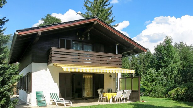 Una casa de dos pisos con un patio cubierto, césped y vistas del bosque circundante.