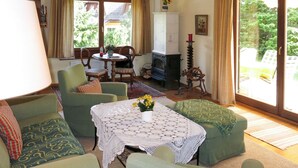 Una sala de estar acogedora con chimenea, muebles tapizados en verde, una mesa con mantel de encaje y grandes ventanales con vistas al exterior.