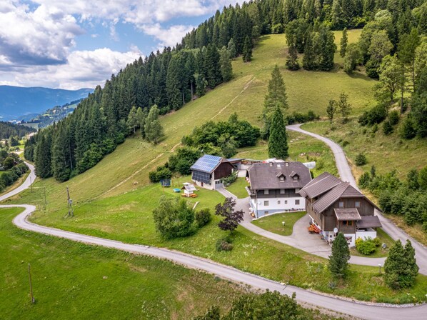 A rural house with a solar panel, surrounded by a forested hillside and a winding road.