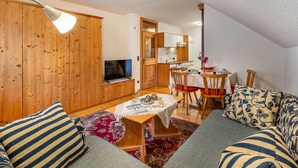 Un salon chaleureux avec un téléviseur, des murs en bois, un coin repas avec une table et des chaises, et une cuisine en arrière-plan.