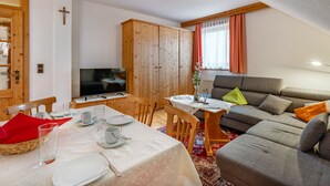 Ein gemütliches Wohnzimmer mit grauem Sofa, einem Fernseher, Holzschränken und einem Essbereich mit Tisch für zwei Personen.