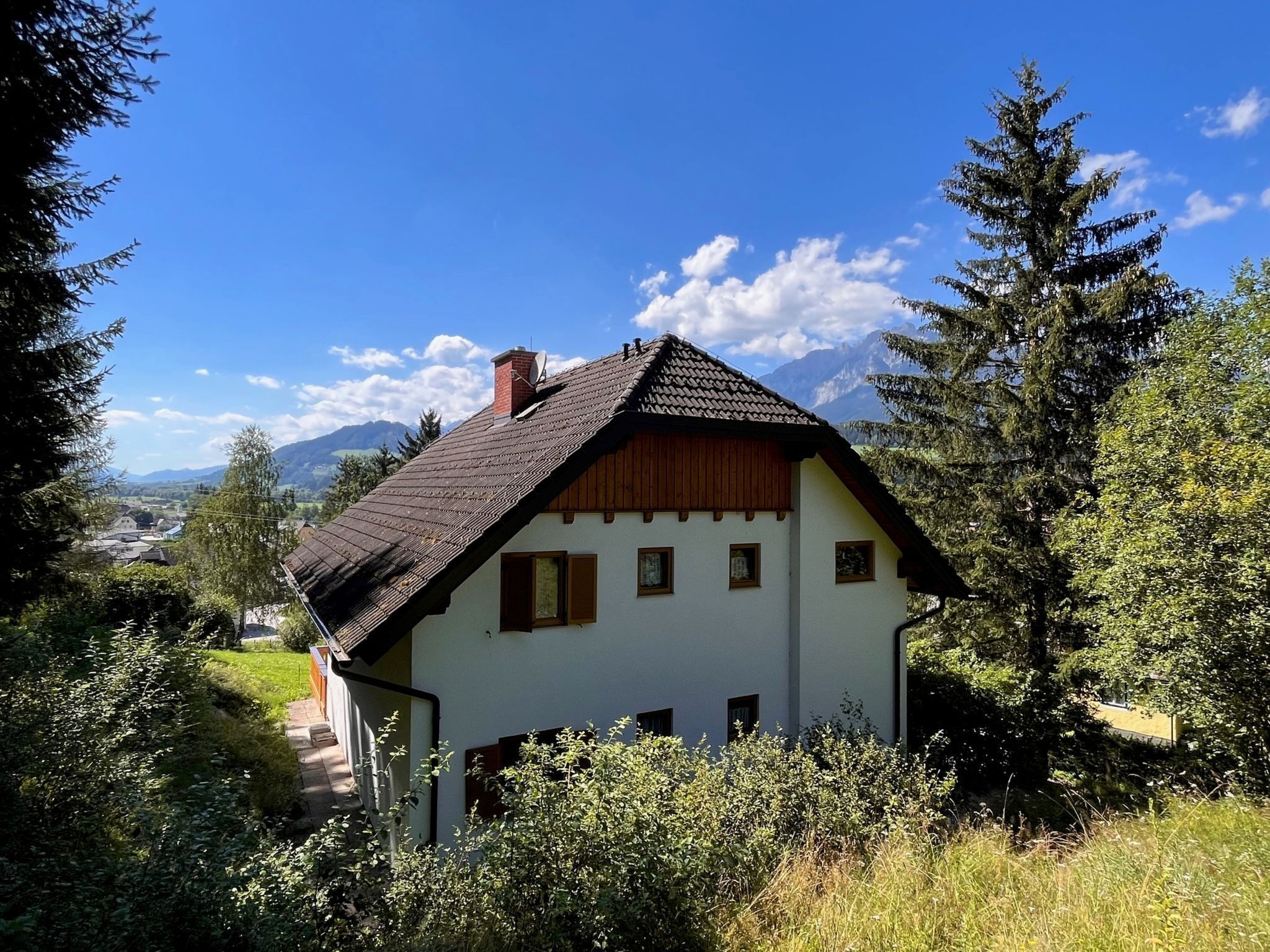 Een klassiek chalet met een schuin dak, omgeven door weelderige begroeiing en bergen op de achtergrond.