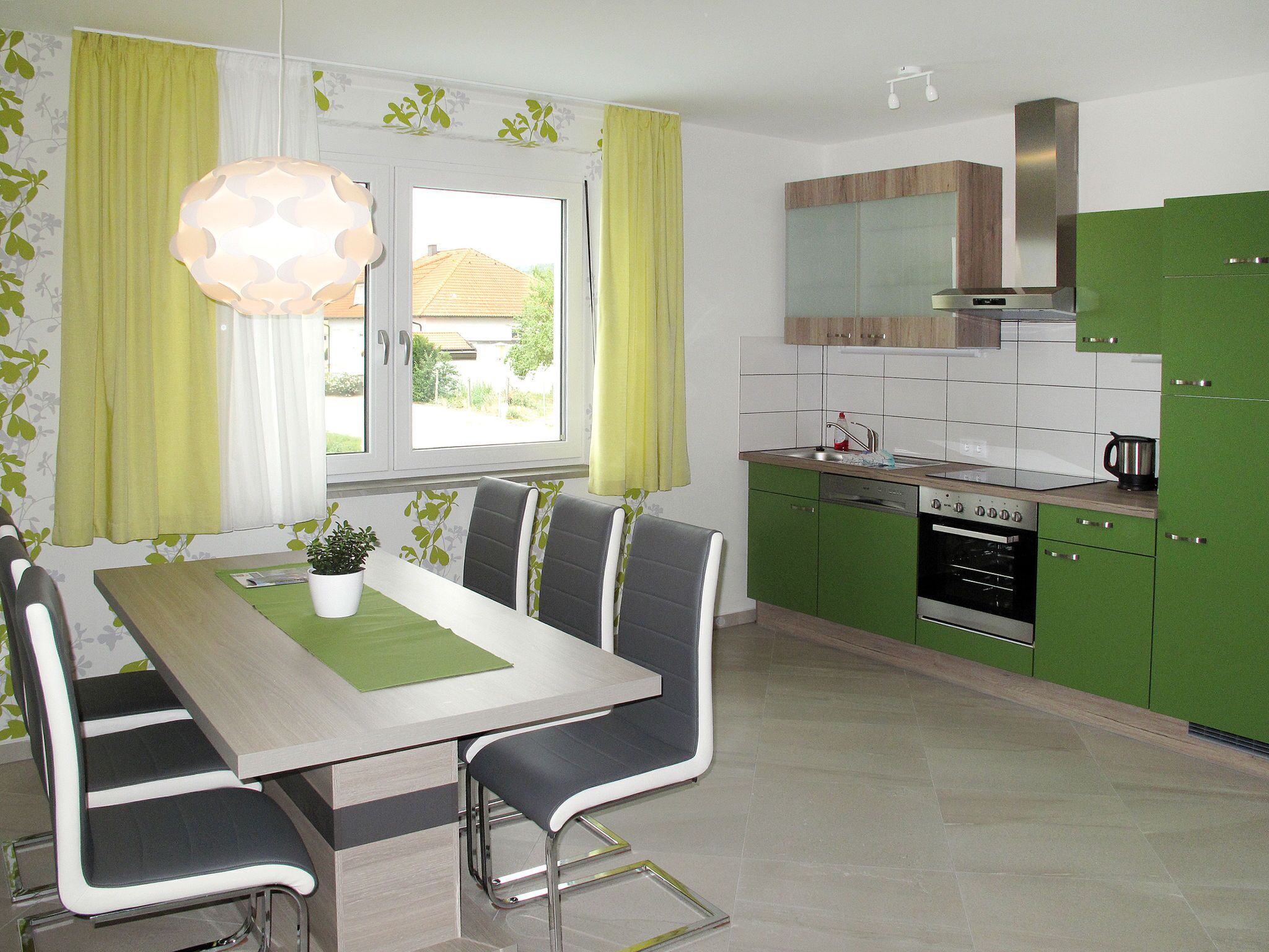 Een moderne keuken met groene kastjes, een roestvrijstalen oven en een eethoek met een tafel en stoelen.