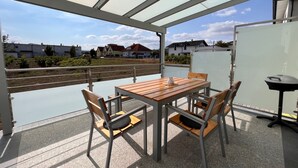 Patio esterno con tavolo e sedie in legno, un barbecue e vista su case sullo sfondo.