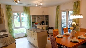 Eine moderne Küche mit Kochinsel, Esstisch aus Holz und Stühlen. Es gibt grüne Vorhänge, ein Fenster mit Aussicht und eine Deckenleuchte.