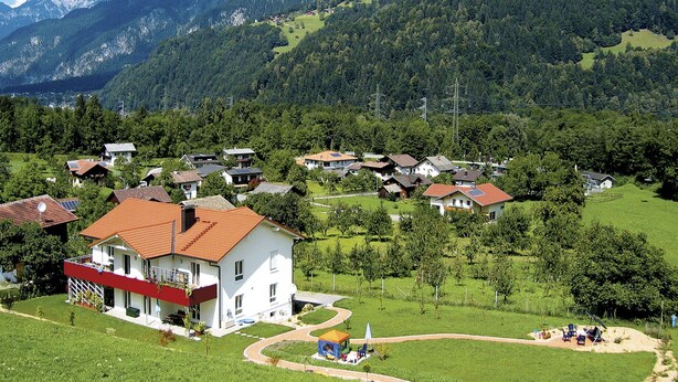 Ein Wohngebiet mit Häusern, einem zentralen weißen Gebäude mit rotem Dach und einer bergigen Kulisse.