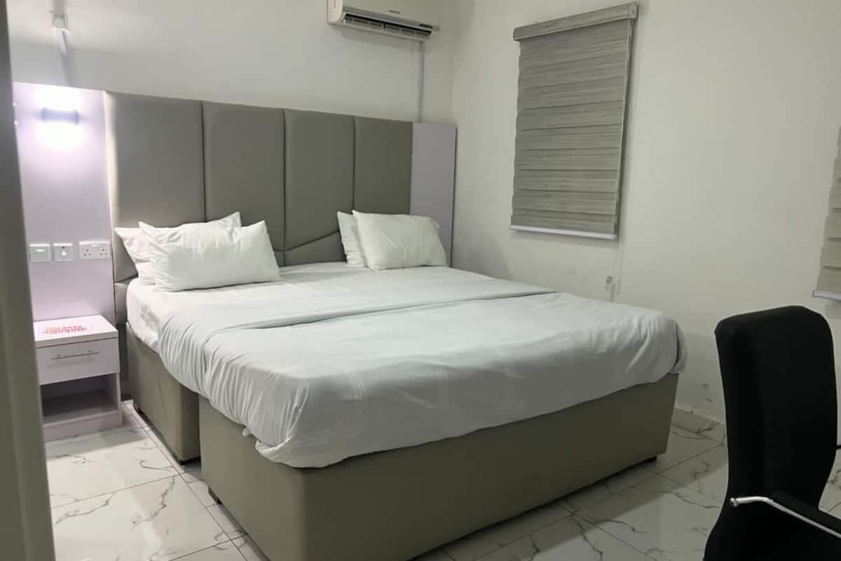 A bedroom with a bed, a nightstand, an air conditioner, a window with blinds, and a chair.