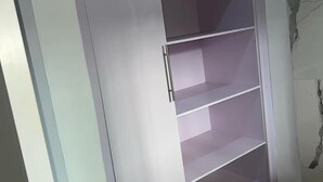 Une armoire blanche, dotée de plusieurs étagères et d’une poignée, se trouve dans une pièce dont le sol est recouvert de carreaux et un des murs est décoré d’un motif imitant le marbre.