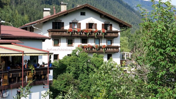 Un bâtiment de style alpin traditionnel, doté d’un balcon orné de fleurs rouges, se dressant sur fond de montagnes verdoyantes.