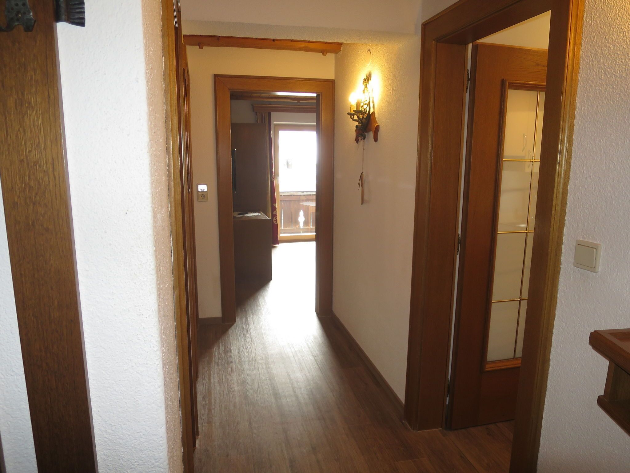 A narrow hallway with wooden flooring and walls, leading to a room with a window and a door.