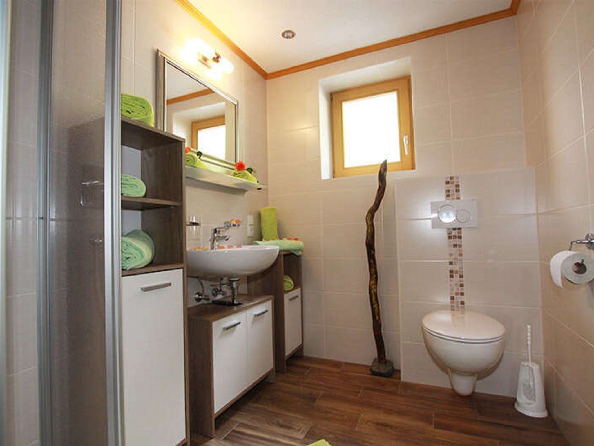 Ein Badezimmer mit Holzboden, Waschbecken in Weiß, Spiegel, Toilette und Duschbereich.