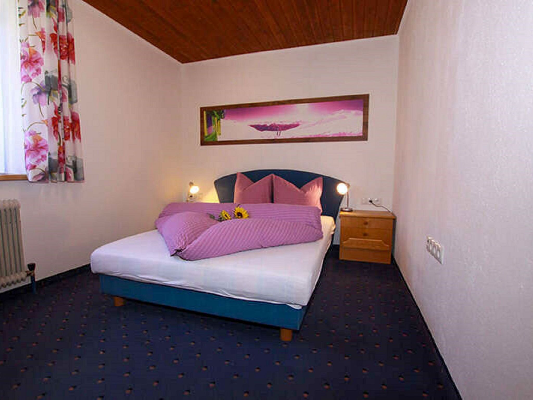 A bedroom with a bed, a nightstand, a lamp, a radiator, and a painting on the wall.