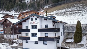 Un immeuble à plusieurs étages doté de balcons, une enseigne indiquant « APART MIRAMONTE », et un paysage enneigé en arrière-plan.