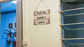 A room with a blue door, a wall-mounted sign that reads "Neste lar habita o amor", and a window with metal blinds.