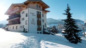 Un edificio de varios pisos con balcones, una pista de esquí al frente y un paisaje montañoso nevado al fondo.