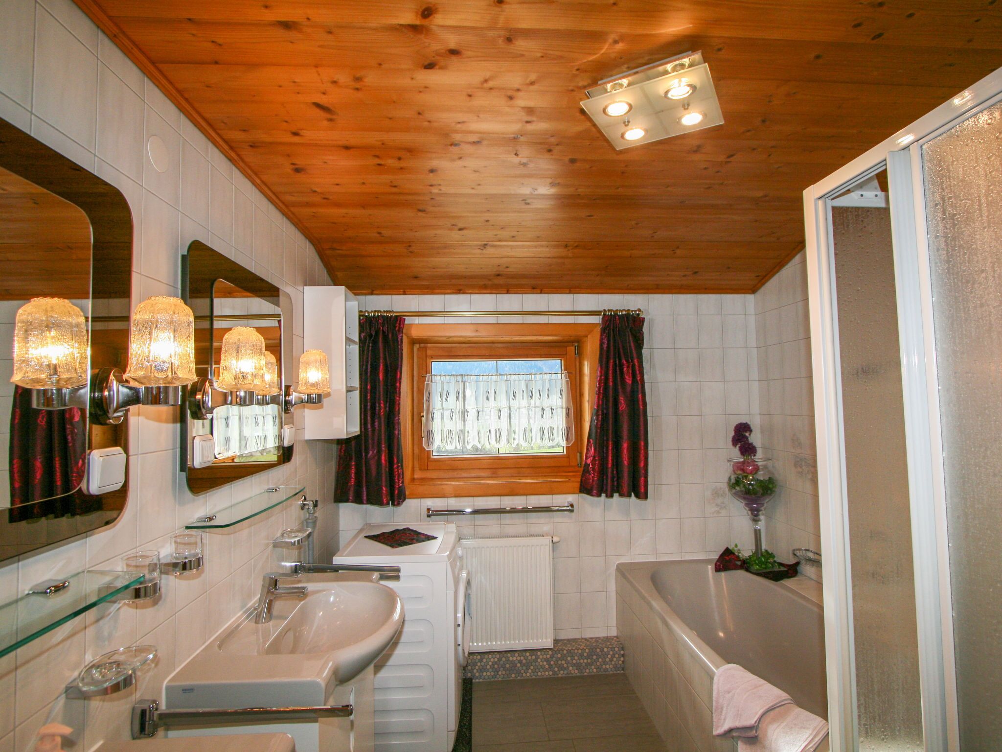 A bathroom with a wooden ceiling, a window with curtains, a bathtub, and a mirror with lights.