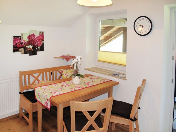 Un coin repas avec une table en bois, deux chaises, un vase de fleurs, une horloge au mur et une fenêtre avec une poutre en bois.