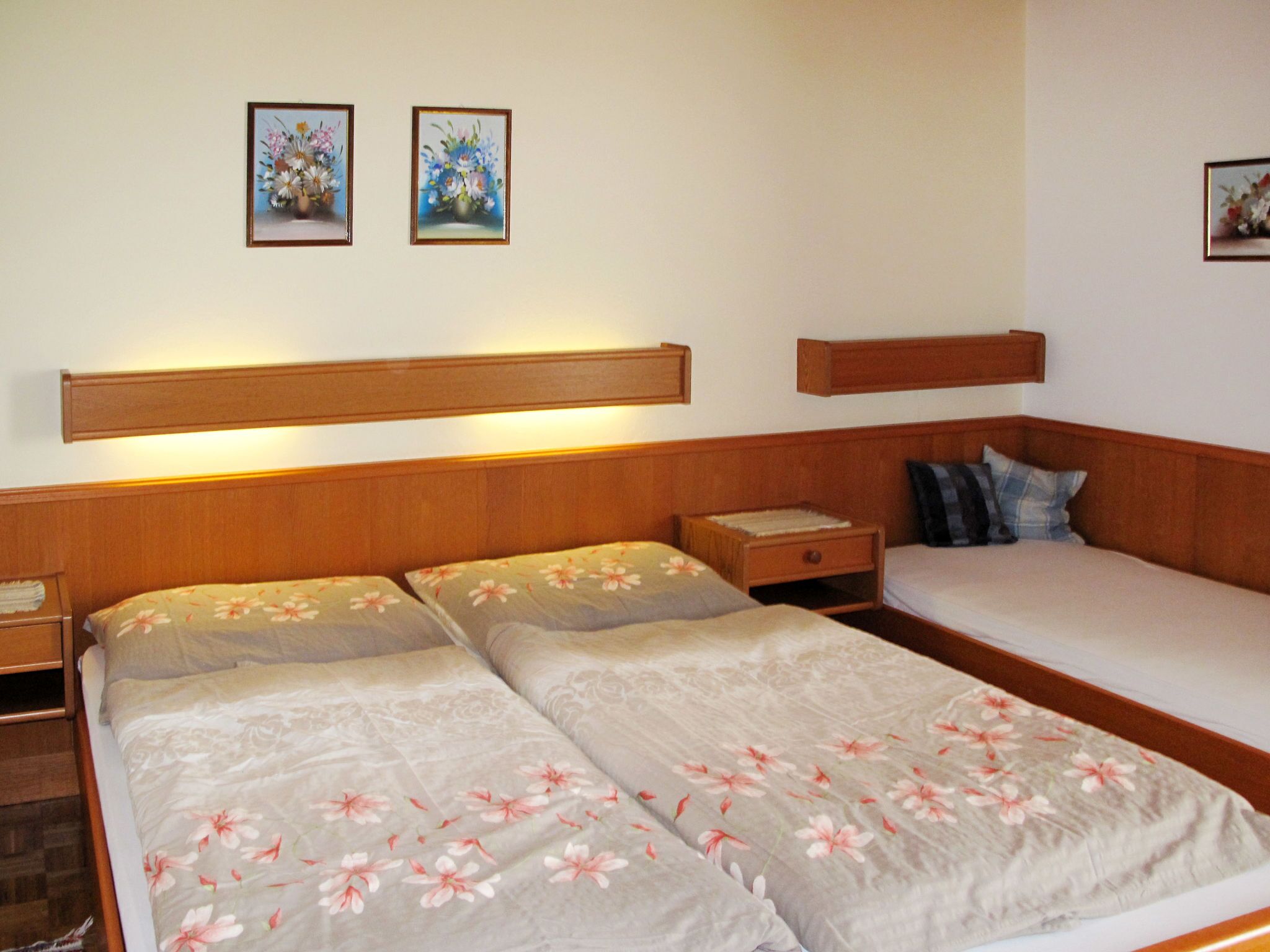 Ein Doppelbett mit floralem Bettbezug, ein Nachttisch, ein Beistelltisch und ein Kopfteil. An der Wand hängen zwei gerahmte Blumenbilder.