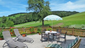 A patio with lounge chairs, a table, and an umbrella, overlooking a green landscape with hills. - Mayrhofer by Interhome (Mondsee)