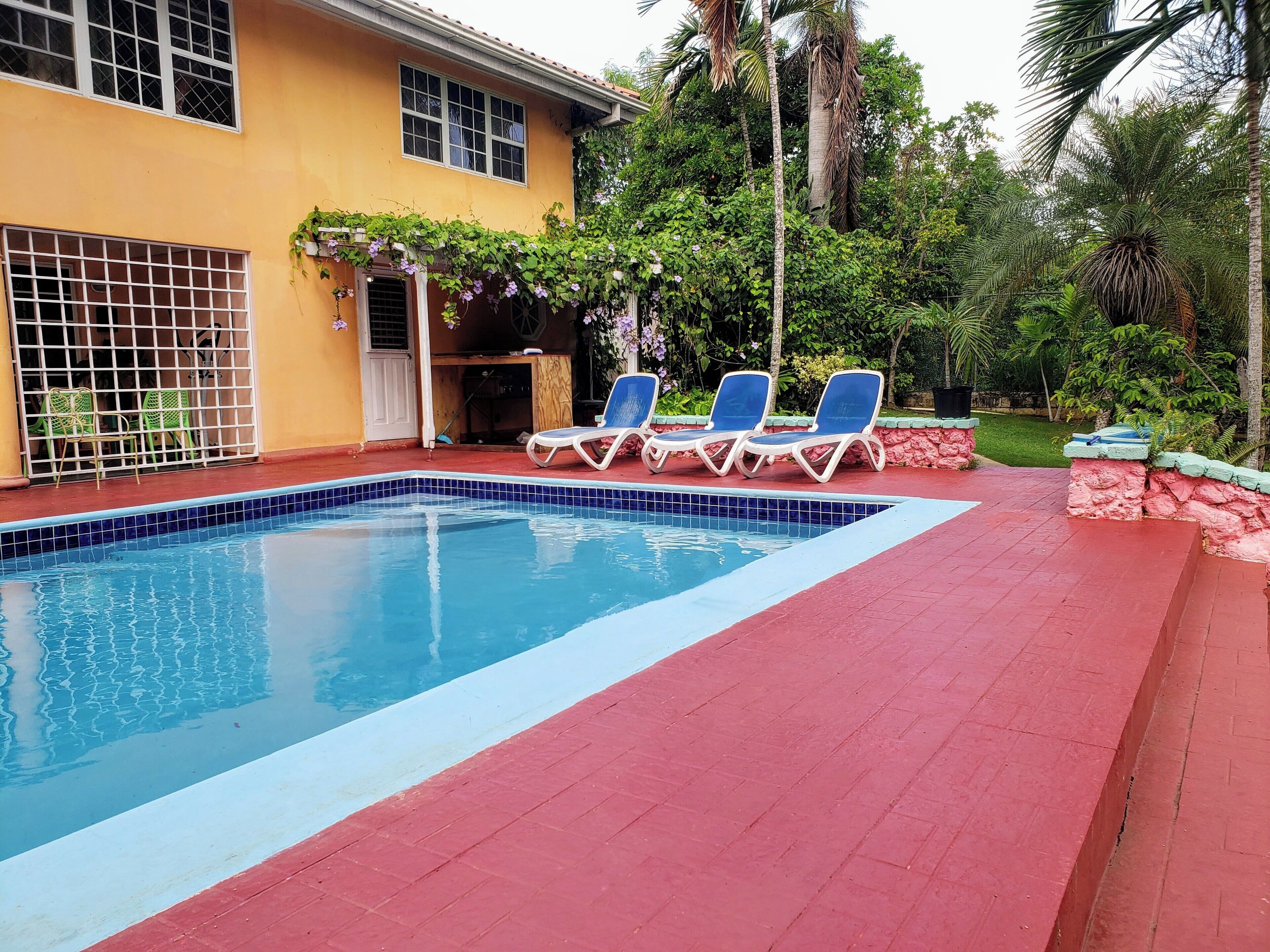 A swimming pool with blue tiles, surrounded by a red deck. There are three lounge chairs and a small covered area with a wooden table and chairs.