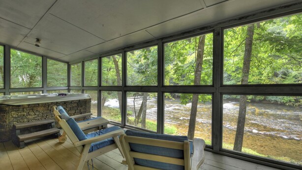 A covered outdoor area with a stone fireplace, wooden lounge chairs, and large windows overlooking a forested area with a stream. - Riverside relaxation and all the comforts of home! - Aska River House (Blue Ridge)