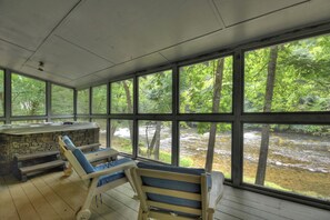 Un espace extérieur abrité doté d’une cheminée en pierre, de fauteuils en bois et de grandes fenêtres donnant sur une zone boisée traversée par un ruisseau.